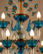 An ornate multi-tier chandelier featuring vibrant turquoise blue glass floral and leaf motifs, clear faceted glass stems, and warm brass-toned metal accents, shown against a neutral beige background.