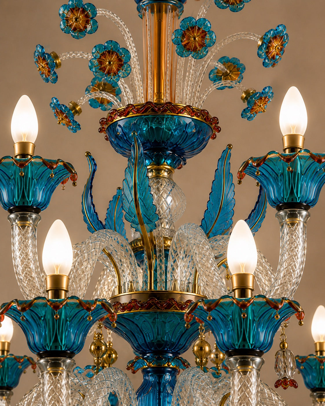 An ornate multi-tier chandelier featuring vibrant turquoise blue glass floral and leaf motifs, clear faceted glass stems, and warm brass-toned metal accents, shown against a neutral beige background.