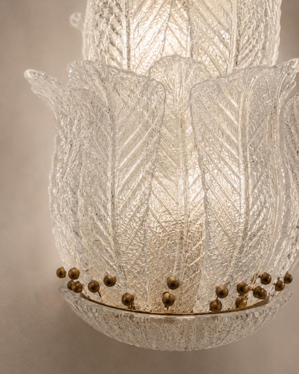 A close-up of a textured clear glass chandelier shade with a leaf-like petal design and a band of small metallic gold beads near the bottom, illuminated from within against a warm neutral background.