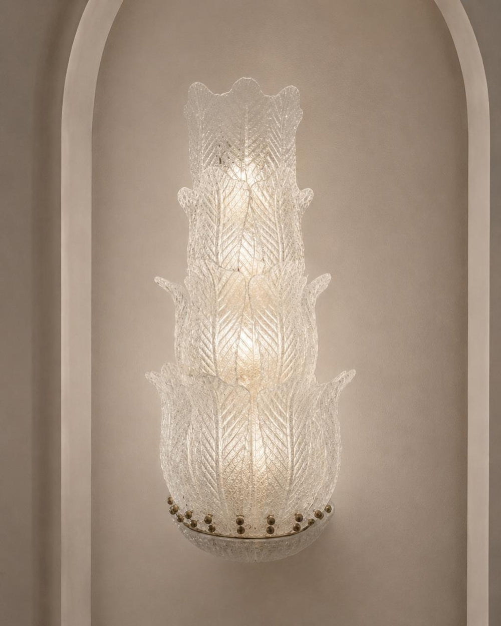 A tiered Murano-style glass wall sconce with a textured, etched leaf pattern, mounted on a beige wall within a white arched niche.