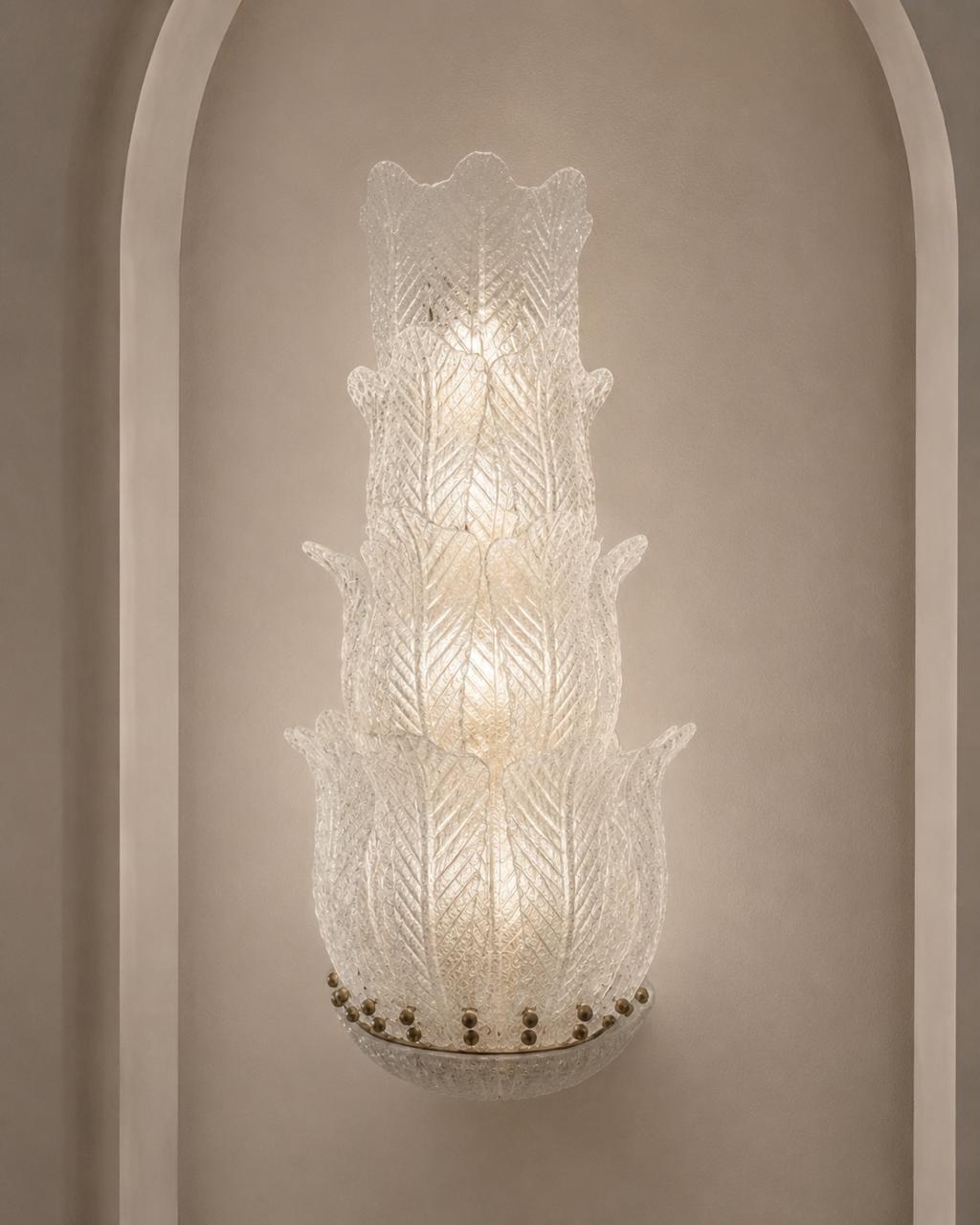 A tiered Murano-style glass wall sconce with a textured, etched leaf pattern, mounted on a beige wall within a white arched niche.