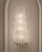 A tiered Murano-style glass wall sconce with a textured, etched leaf pattern, mounted on a beige wall within a white arched niche.