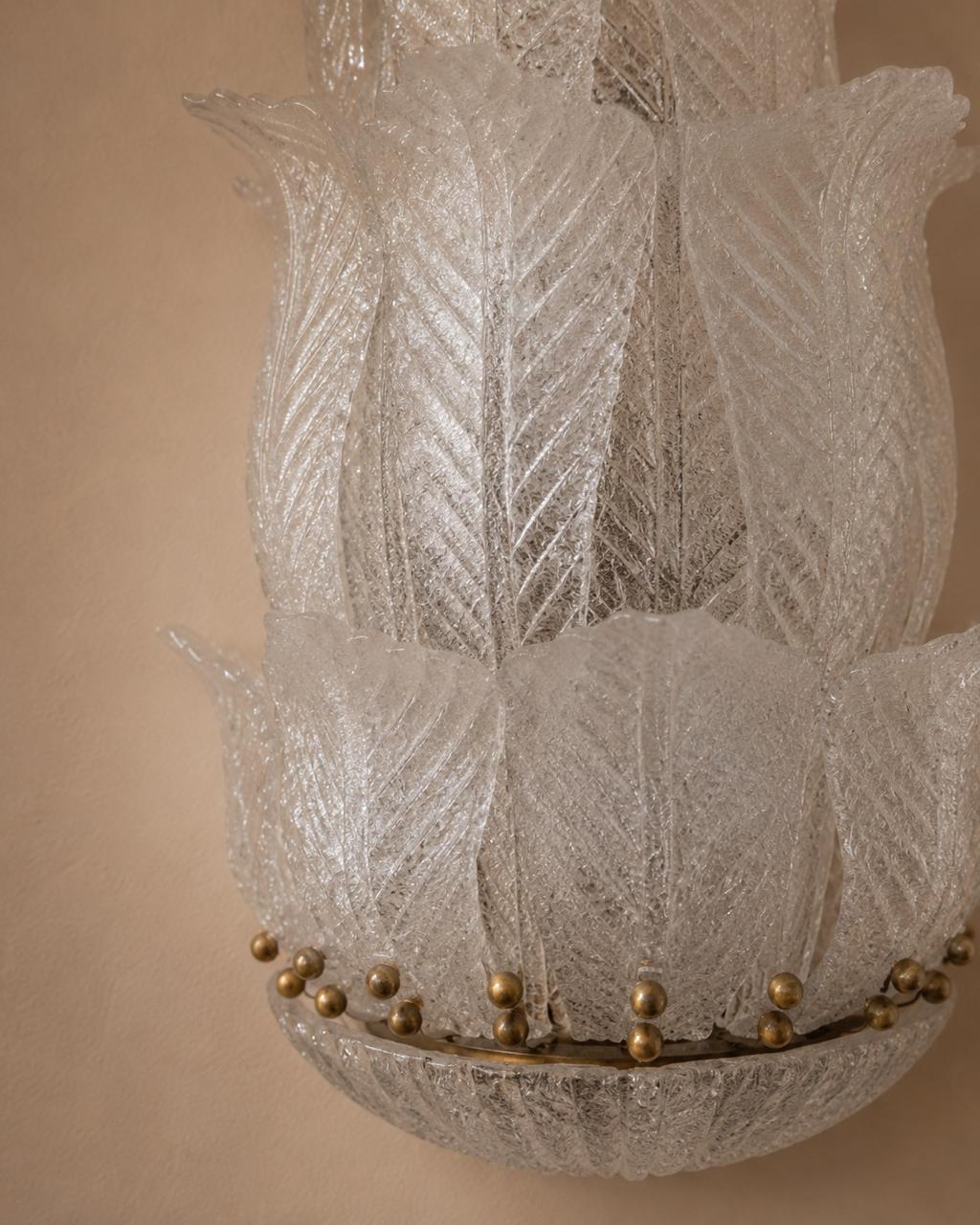 A close-up of a textured glass chandelier shade with layered, lotus-like petal shapes and small gold metallic beads along the rim, set against a warm beige background.