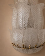 A close-up of a textured glass chandelier shade with layered, lotus-like petal shapes and small gold metallic beads along the rim, set against a warm beige background.