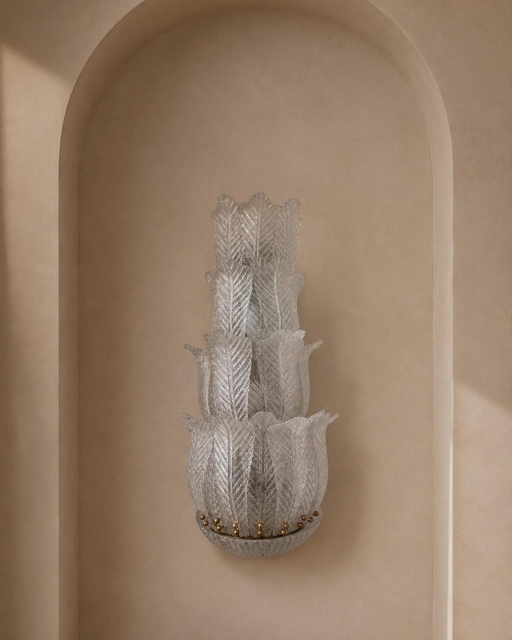 A sculptural, multi-tiered wall sconce made of textured, translucent white glass with a leaf-like pattern, mounted on a beige plaster wall in a recessed niche.