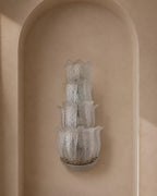 A sculptural, multi-tiered wall sconce made of textured, translucent white glass with a leaf-like pattern, mounted on a beige plaster wall in a recessed niche.