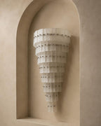 A tiered, cone-shaped wall sconce made of translucent white pleated material, mounted on a beige plaster wall within a recessed arch.