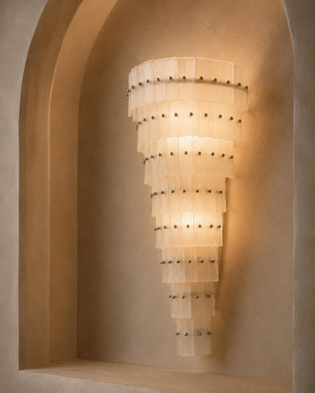 A tiered wall sconce with translucent rectangular glass shades and small metallic studs, mounted on a warm beige plaster wall within an arched niche.