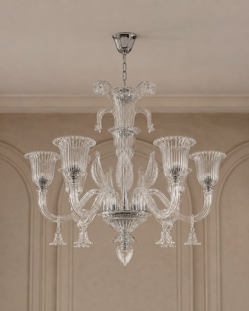 A clear glass chandelier with six fluted candle-style arms and a central stem, hanging from a silver ceiling canopy in an indoor room with beige walls and molding.