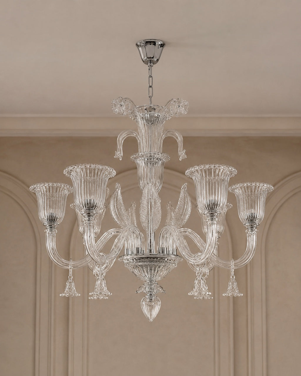 A clear glass chandelier with six fluted candle-style arms and a central stem, hanging from a silver ceiling canopy in an indoor room with beige walls and molding.