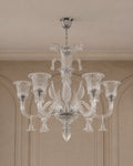 A clear glass chandelier with six fluted candle-style arms and a central stem, hanging from a silver ceiling canopy in an indoor room with beige walls and molding.