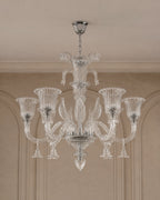 A clear glass chandelier with six fluted candle-style arms and a central stem, hanging from a silver ceiling canopy in an indoor room with beige walls and molding.