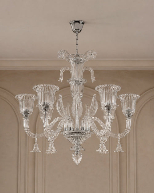 A clear glass chandelier with six fluted candle-style arms and a central stem, hanging from a silver ceiling canopy in an indoor room with beige walls and molding.
