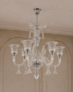 A multi-arm crystal chandelier with a polished silver-toned metal frame and clear glass shades, hanging from a ceiling in an elegant interior with beige walls and molding.