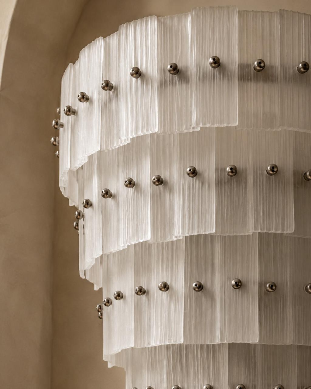 A close-up of a modern cylindrical chandelier with layered, translucent white textured panels and evenly spaced metallic silver round studs, set against a warm beige background.