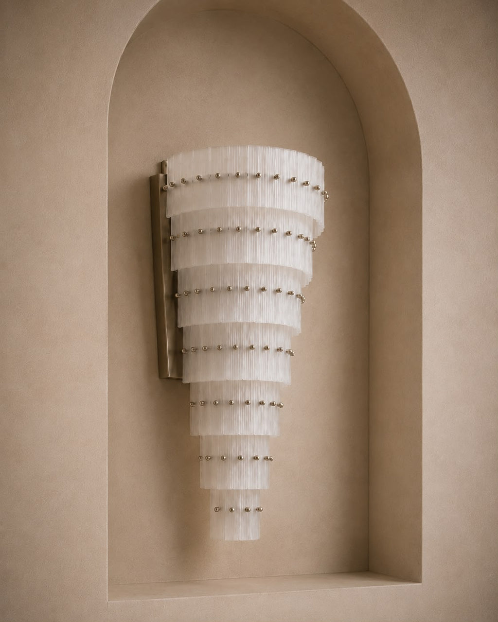A modern wall sconce with a tapered, tiered design made of translucent white pleated glass discs, accented with small metallic studs, mounted on a beige plaster wall in a recessed niche.