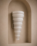 A modern wall sconce with a tapered, tiered design made of translucent white pleated glass discs, accented with small metallic studs, mounted on a beige plaster wall in a recessed niche.