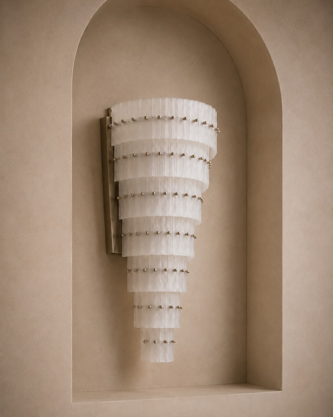 A modern wall sconce with a tapered, tiered design made of translucent white pleated glass discs, accented with small metallic studs, mounted on a beige plaster wall in a recessed niche.