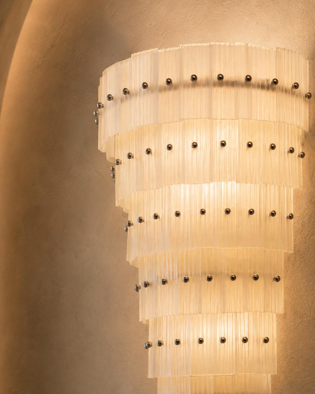A modern spiral chandelier with translucent pleated glass or acrylic rings and evenly spaced dark metal studs, illuminated against a warm beige plaster wall.