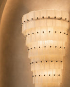 A modern spiral chandelier with translucent pleated glass or acrylic rings and evenly spaced dark metal studs, illuminated against a warm beige plaster wall.
