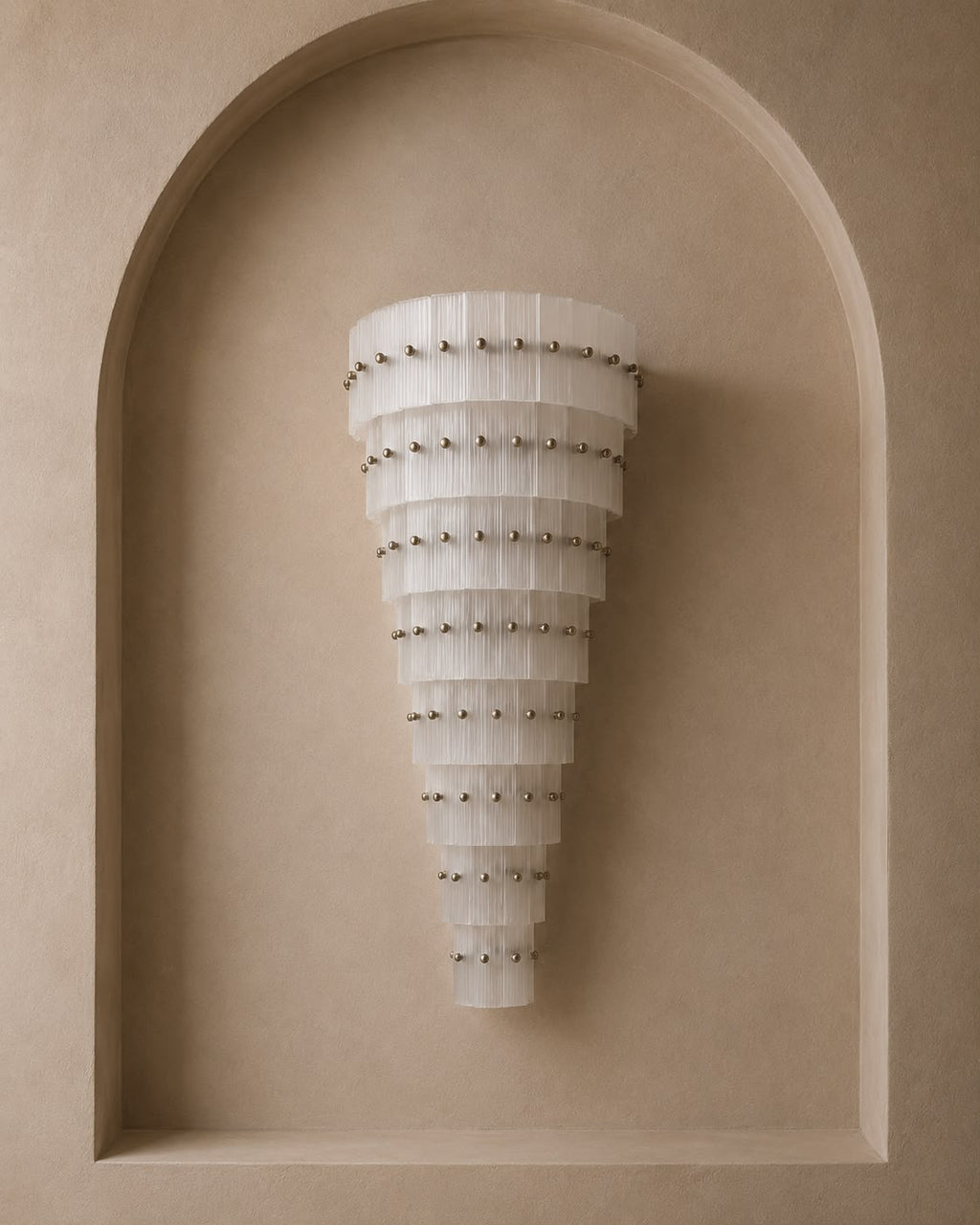 A modern tiered wall sconce with translucent white curved panels and small metallic studs, mounted on a beige plaster wall within an arched recess.