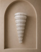 A modern tiered wall sconce with translucent white curved panels and small metallic studs, mounted on a beige plaster wall within an arched recess.
