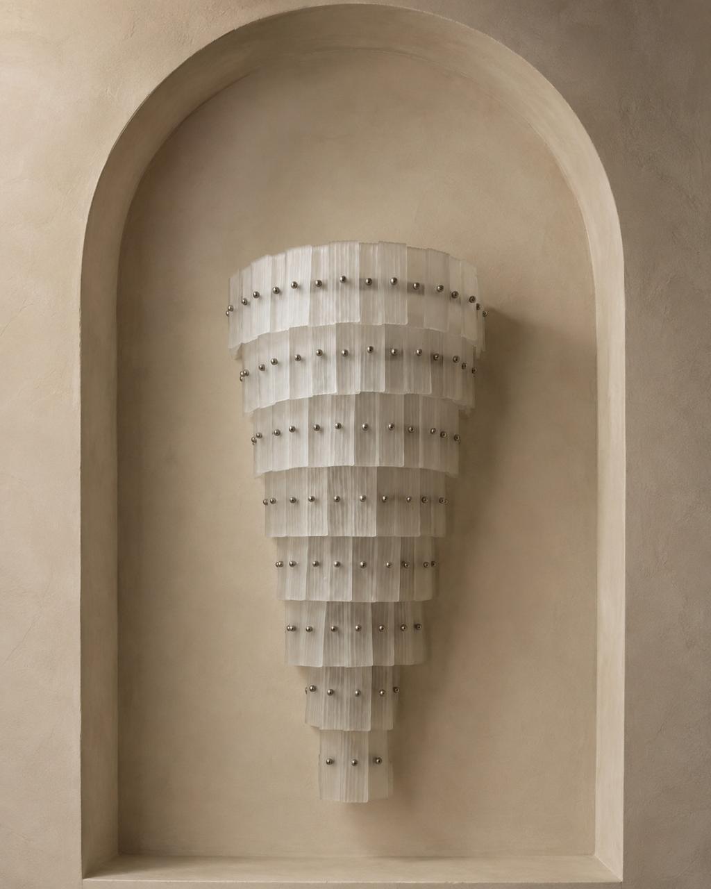 A modern wall sconce with a tiered, cone-shaped design made of translucent white pleated panels and accented with small silver studs, mounted on a beige plaster wall within a recessed arch.