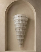 A modern wall sconce with a tiered, cone-shaped design made of translucent white pleated panels and accented with small silver studs, mounted on a beige plaster wall within a recessed arch.