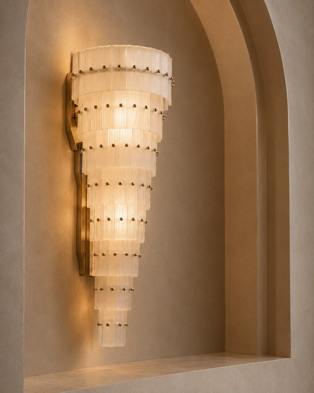 A tiered wall sconce with a warm brass finish and translucent white glass shades, mounted on a beige plaster wall in an arched niche.