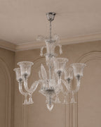 A clear glass chandelier with multiple fluted candle-style arms and hanging crystal drops, suspended from a silver ceiling canopy in an elegant beige room with decorative wall molding.