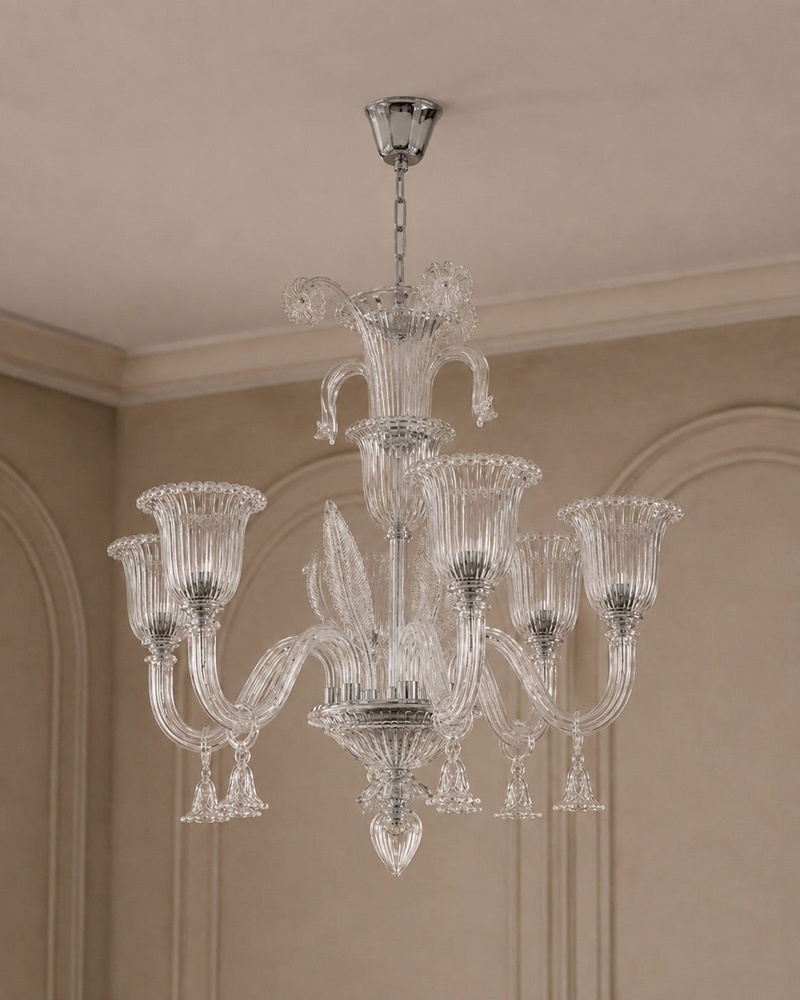 A clear glass chandelier with multiple fluted candle-style arms and hanging crystal drops, suspended from a silver ceiling canopy in an elegant beige room with decorative wall molding.