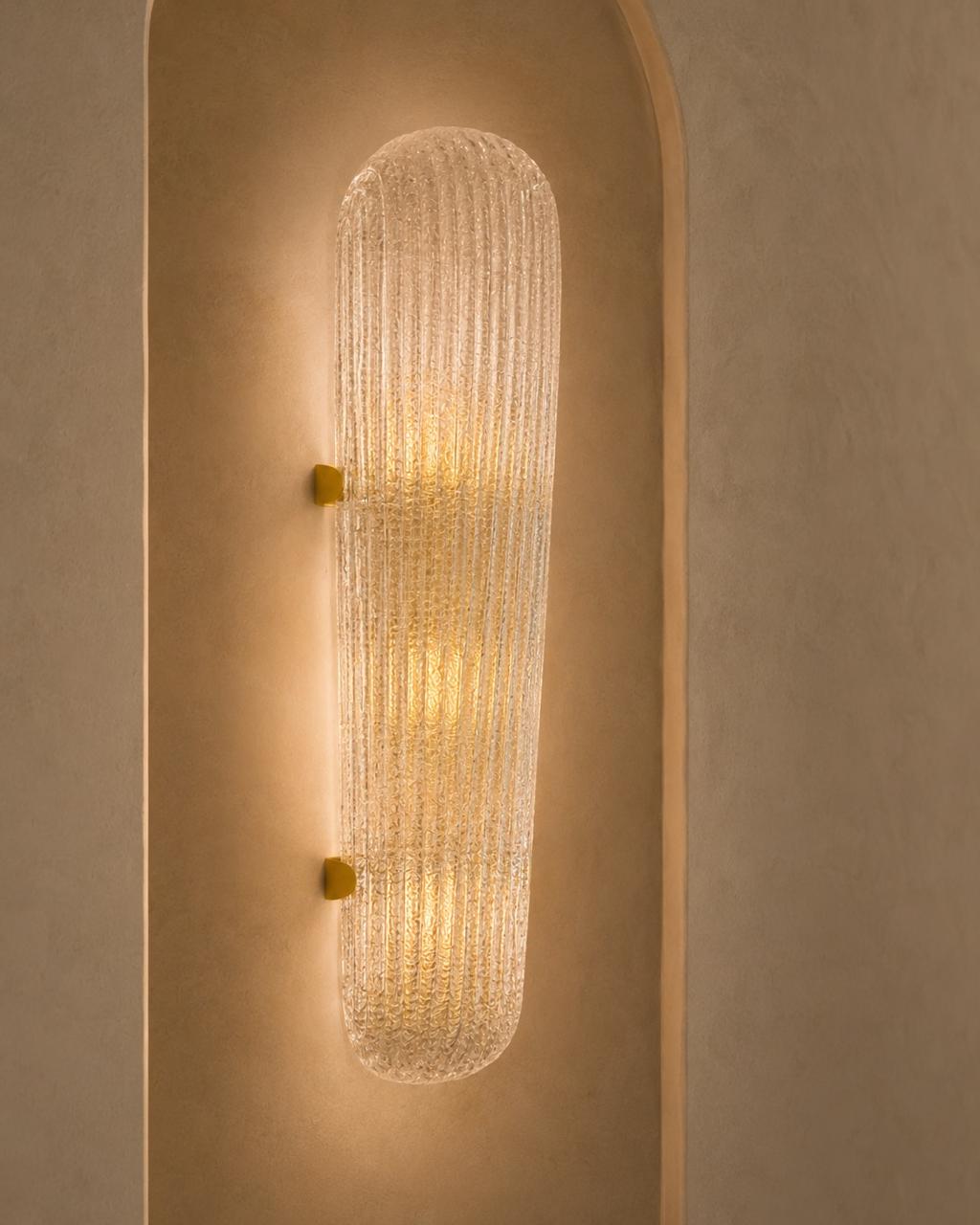 A modern wall sconce with a ribbed, textured clear glass shade and small gold mounting brackets, mounted on a warm beige plaster wall.