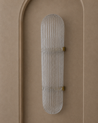 A modern vertical wall sconce with a ribbed clear glass shade and small brass hardware, mounted on a warm beige textured wall with an arched recess.