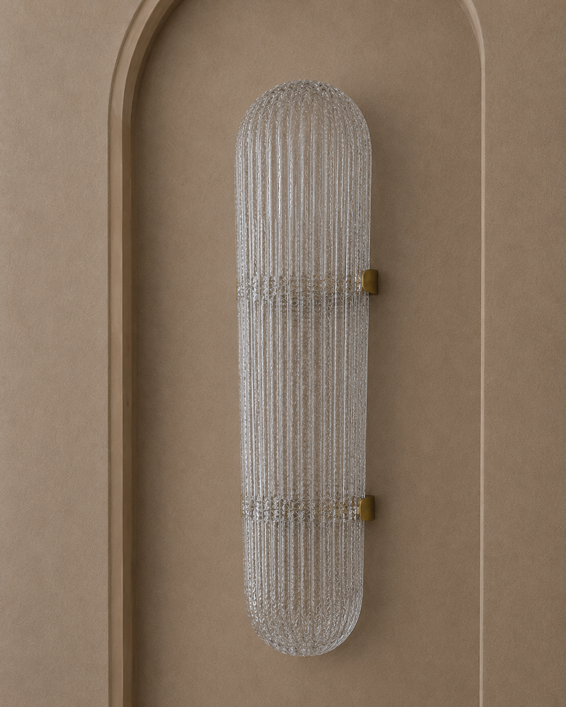 A modern vertical wall sconce with a ribbed clear glass shade and small brass hardware, mounted on a warm beige textured wall with an arched recess.