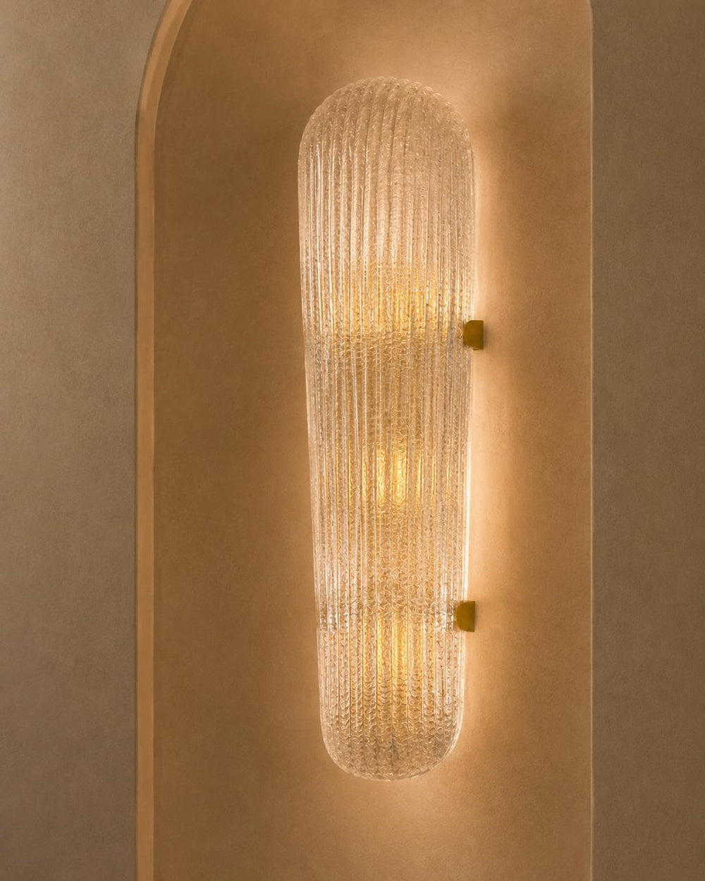 A modern cylindrical wall sconce with a ribbed, textured clear glass shade and warm yellow light, mounted on a beige plaster wall with an arched niche.