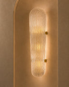 A modern cylindrical wall sconce with a ribbed, textured clear glass shade and warm yellow light, mounted on a beige plaster wall with an arched niche.