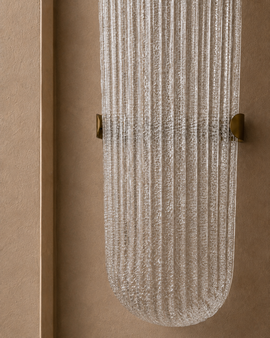 A modern wall-mounted light fixture with a ribbed, textured clear glass shade and brass hardware, shown against a warm beige stucco wall.
