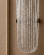 A modern wall-mounted light fixture with a ribbed, textured clear glass shade and brass hardware, shown against a warm beige stucco wall.