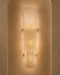 A modern wall sconce with a ribbed, textured glass shade that glows with warm light, mounted on a textured beige plaster wall.