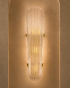 A modern wall sconce with a ribbed, textured glass shade that glows with warm light, mounted on a textured beige plaster wall.