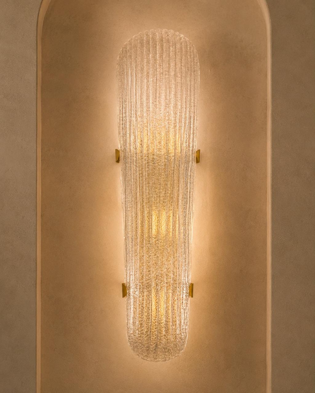 A modern wall sconce with a ribbed, textured glass shade that glows with warm light, mounted on a textured beige plaster wall.