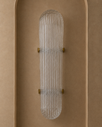 A modern wall-mounted light fixture with a ribbed clear glass shade and small gold metal brackets, centered against a warm beige textured wall.