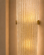 A modern wall sconce featuring a textured, ribbed clear glass cylinder that glows with a warm golden light, mounted on a beige textured wall with two small gold metal brackets on either side.
