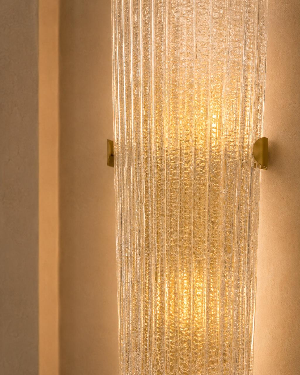 A modern wall sconce featuring a textured, ribbed clear glass cylinder that glows with a warm golden light, mounted on a beige textured wall with two small gold metal brackets on either side.