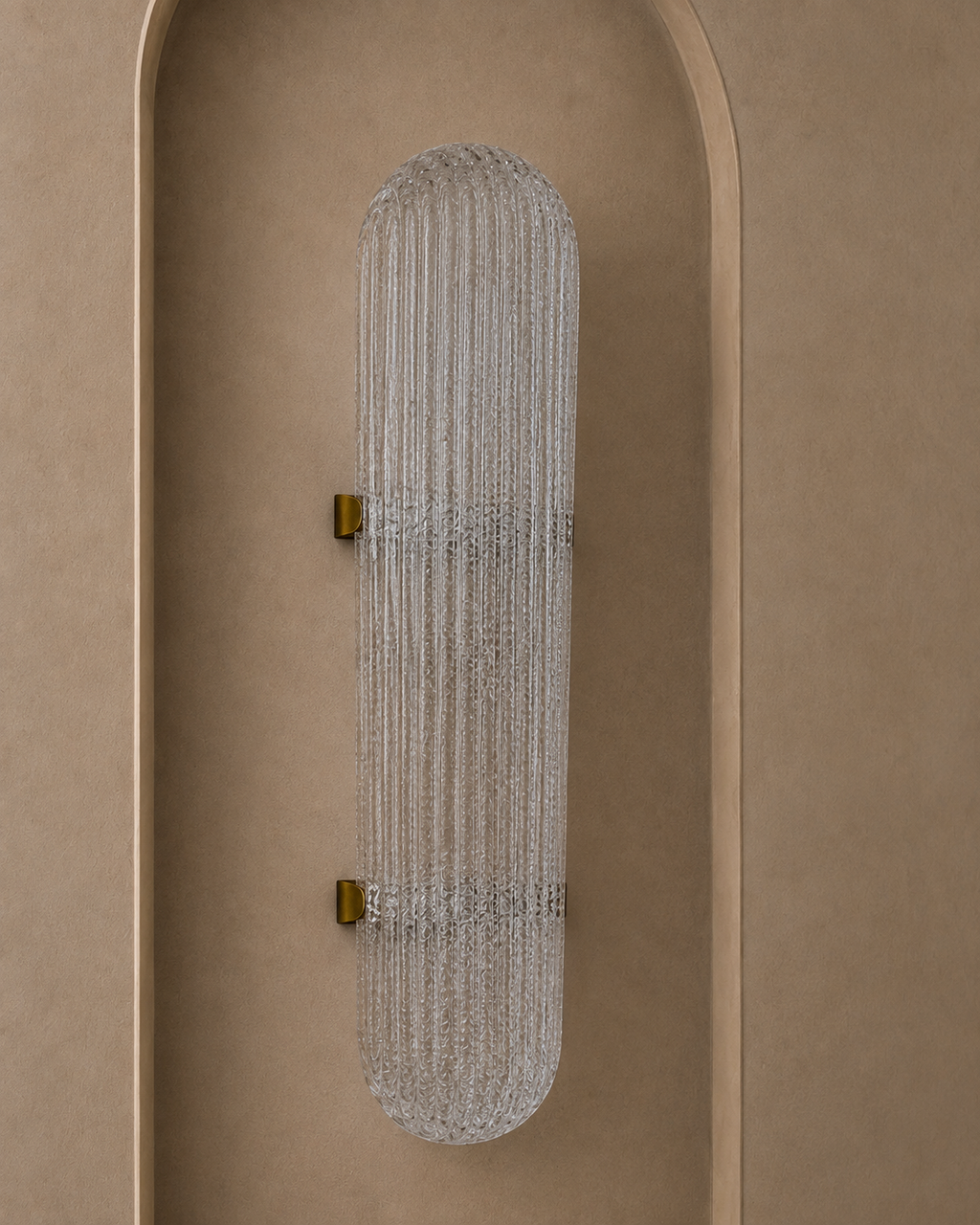 A modern wall sconce with a ribbed, vertically oriented cylindrical glass shade and small gold wall mounts, shown installed on a warm beige stucco wall with an arched recess.