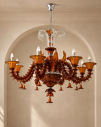 An ornate multi-arm chandelier made of amber-brown translucent glass with sculpted details, clear glass accents, and multiple candle-style bulbs, hanging in front of a beige arched wall.