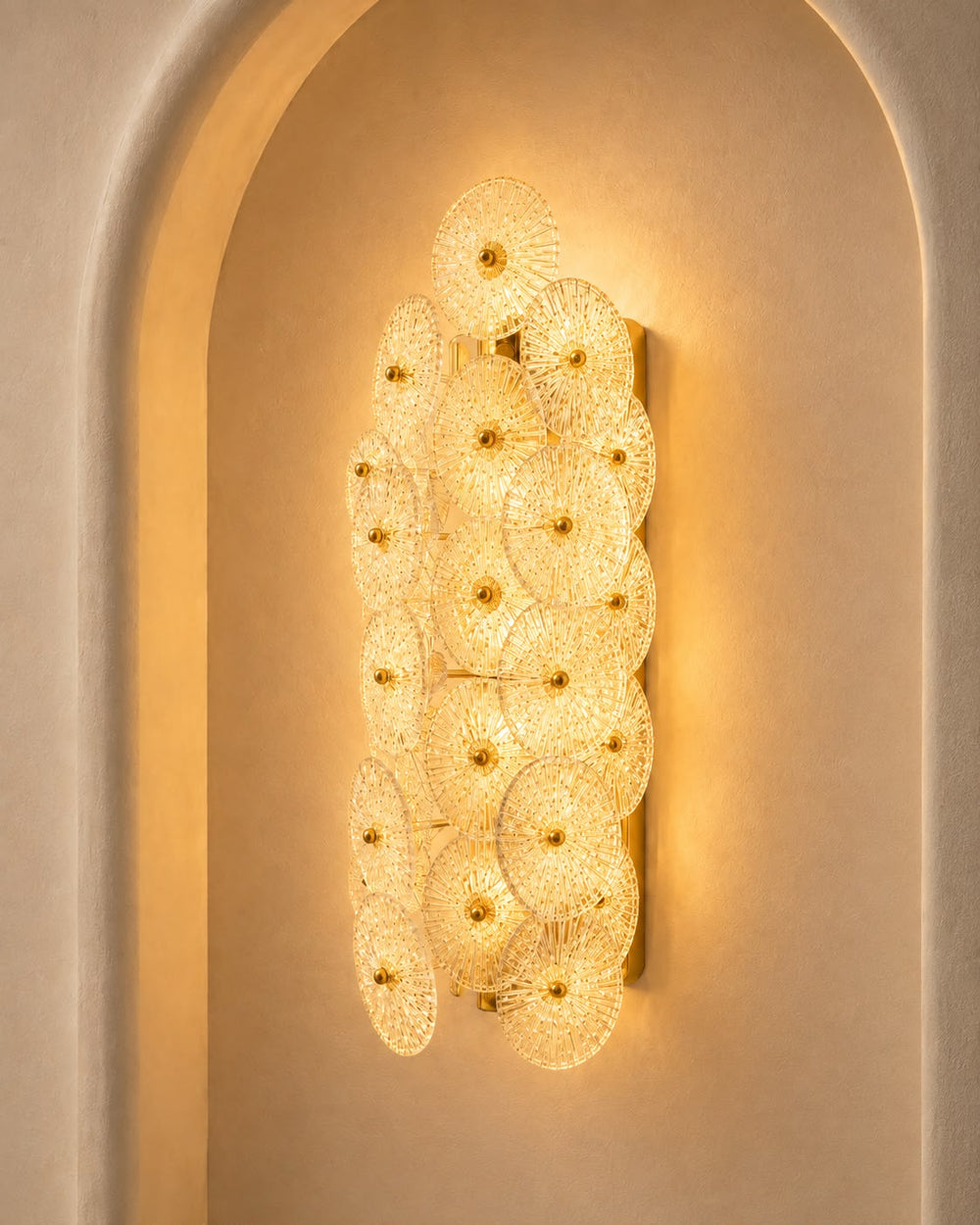A vertical wall sconce featuring multiple overlapping circular glass shades with gold-toned centers, mounted on a beige plaster wall within an arched recess.