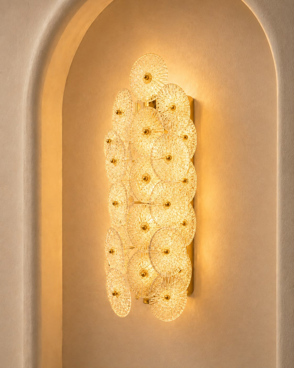 A vertical wall sconce featuring multiple overlapping circular glass shades with gold-toned centers, mounted on a beige plaster wall within an arched recess.