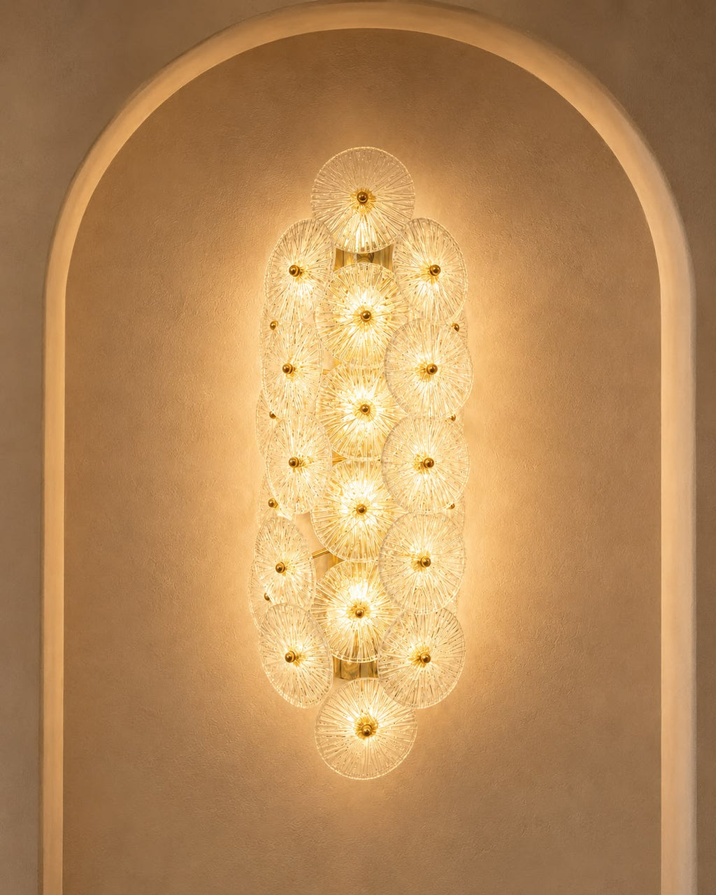 A modern wall sconce featuring multiple overlapping circular glass shades with gold-toned metal frames, mounted on a warm beige plaster wall within an arched niche.