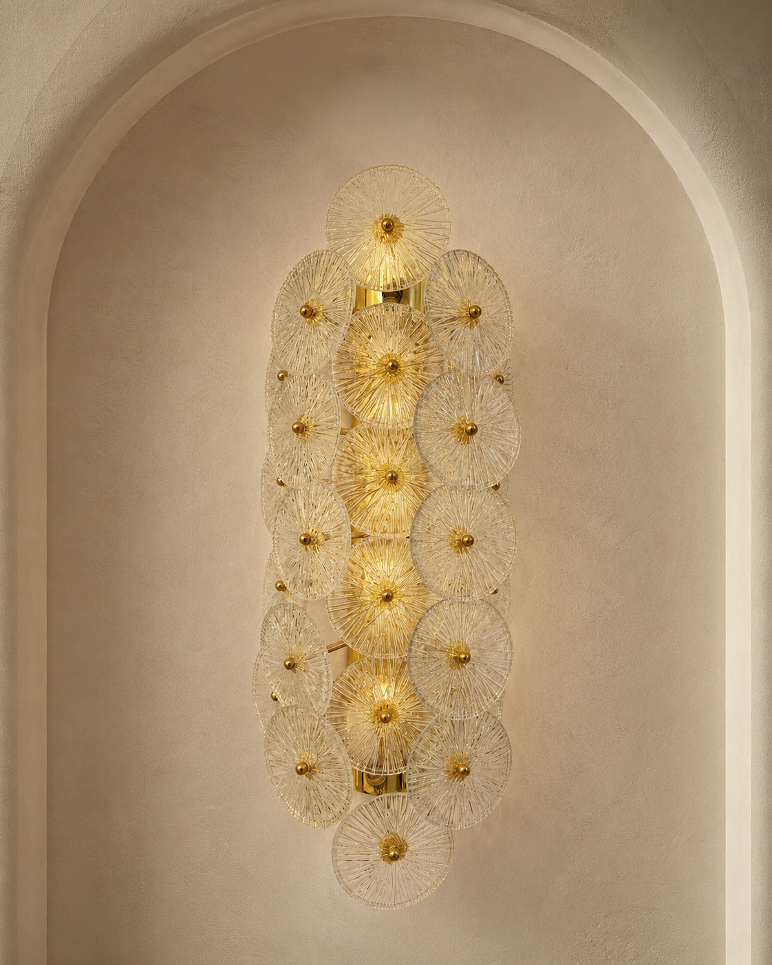 A vertical wall sconce with multiple overlapping circular glass shades and gold-toned metal accents, mounted in a tall arched alcove against a warm beige plaster wall.